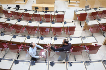 | Foto: Stefan Obermeier Vogelperspektive. Zwei Mitarbeiter halten eine durchsichtige Trennscheibe im Plenarsaal in den Händen.